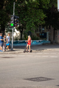 Фотография женщины на самокате с татуировкой на ноге, пересекающей улицу на фоне городского пейзажа.