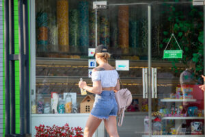 Фотография девушки в кепке, белой майке и джинсовых шортах с татуировкой крыла на плече, стоящей у витрины магазина.