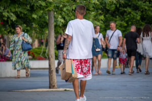 Фото тату на руке мужчины, идущего по улице. Татуировка выполнена в темных тонах и занимает большую часть предплечья. Мужчина одет в белую футболку и красные шорты.
