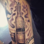Фото тату бутылки Belvedere Vodka на предплечье, выполненное в черно-серых тонах с добавлением красных брызг краски, создающих динамичный эффект.