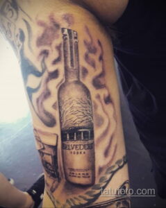 Фото тату бутылки Belvedere Vodka на предплечье, выполненное в черно-серых тонах с добавлением красных брызг краски, создающих динамичный эффект.