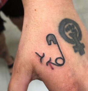 Фото тату булавки на руке с надписью 'XO' и символом феминизма. Черно-белый рисунок татуировки, сочетание символов и текста.