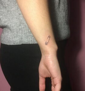 Фото тату маленькой булавки на запястье. Черно-белый рисунок татуировки в стиле минимализм, четкие линии, небольшие размеры.