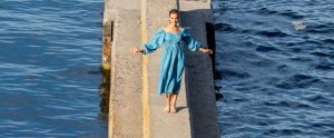 Фотография женщины в голубом платье, стоящей у воды с распущенными руками. На руке мужчины видна татуировка, частично скрытая одеждой.