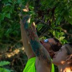 Фотография руки с татуировкой, тянущейся к ветке дерева. Тату выполнено в акварельной технике с использованием ярких цветов.