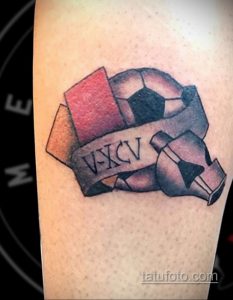 Фото тату футбольного мяча с римскими цифрами 'V-XCV' на ленте. Цветная татуировка, символизирующая дату или важную цифру.