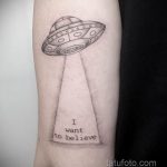 Фото минималистичной татуировки на предплечье: изображение НЛО и надпись 'I want to believe', выполненные тонкими линиями в черно-белой гамме.
