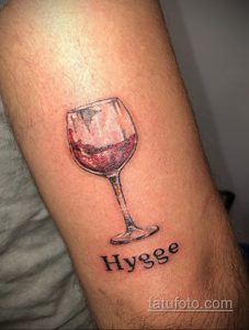 Фото тату: реалистичный рисунок бокала красного вина с надписью "Hygge". Небольшая татуировка на руке.