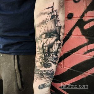 Фото тату: реалистичный рисунок парусника на предплечье, выполненный в черно-серых тонах с детализированными волнами и облаками.