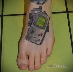 Фото тату Game Boy на стопе. Черно-серый рисунок татуировки с элементами пиксель-арта и яркими красными акцентами. Идеально для поклонников ретро-игр.