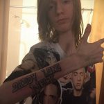 Фотография женщины с татуировкой на руке с надписью "Daddy Long Neck". На фоне видна футболка с изображением двух людей.
