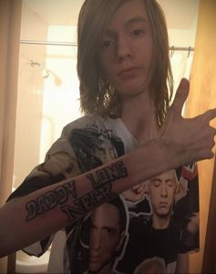 Фотография женщины с татуировкой на руке с надписью "Daddy Long Neck". На фоне видна футболка с изображением двух людей.