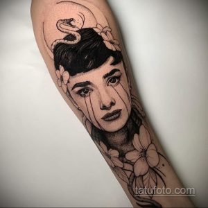 Черно-белая татуировка с портретом плачущей Одри Хепберн, обвитой змеей и украшенной цветами. Детализированный рисунок, готический стиль.