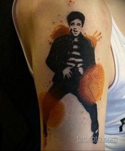 Фото тату Элвиса Пресли на плече, выполненное в акварельной технике с использованием черного и коричневых оттенков. Размытые края и эффект брызг.
