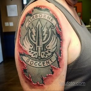 Фото татуировки с изображением рваного флага с эмблемой SOCCENT и надписью 'AIRBORNE' на плече. Черно-серая татуировка в реалистичном стиле.