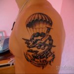 Фото тату на плече: самолет с раскрытым парашютом и силуэтами парашютистов, выполненное в черно-серых тонах, детальный рисунок татуировки.