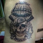 Фото тату черепа в парашюте с горящими крыльями на руке. Черно-серый рисунок с агрессивным дизайном и надписью. Идеи для тату, символизирующие смелость и отвагу.