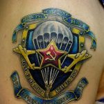 Фото тату на плече с изображением десантного щита с парашютом, звездой и надписями, включая даты и аббревиатуры. Татуировка выполнена в ярких цветах.