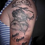 Фото тату на руке с изображением вертолета, из которого выпрыгивают парашютисты. Татуировка выполнена в черно-серых тонах.