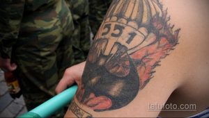Черно-красная татуировка на руке с изображением черепа под раскрытым парашютом. Внизу надпись и элементы декора.