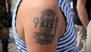 Фото тату парашютиста под раскрытым куполом на плече, выполненное в черно-серых тонах. Детализированный рисунок татуировки с надписями.