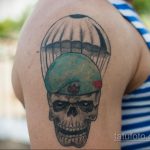 Фото татуировки на плече с изображением черепа в зеленом берете. Тату выполнено в черно-серых и зеленых тонах, выглядит потертым.