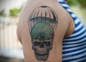 Фото татуировки на плече с изображением черепа в зеленом берете. Тату выполнено в черно-серых и зеленых тонах, выглядит потертым.
