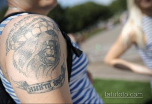 Фото татуировки на плече с изображением черепа в кокарде. Тату выполнено в черно-серых тонах, выглядит выцветшим и потертым.