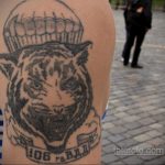 Фото татуировки на плече с изображением головы тигра в парашютном шлеме. Тату выполнено в черно-серых тонах с использованием штриховки.