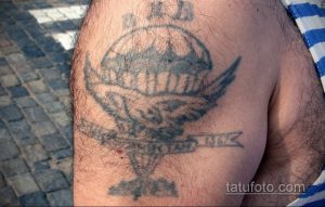 Фото старой татуировки на плече с изображением черепа в окружении надписи. Тату выполнено в черно-серых тонах, выглядит выцветшим и потертым.