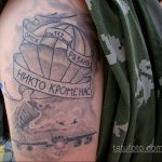Фото татуировки на плече: изображение раскрытого парашюта, самолета и надписи, выполненное в черно-серых тонах.