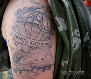 Фото татуировки на плече: изображение раскрытого парашюта, самолета и надписи, выполненное в черно-серых тонах.