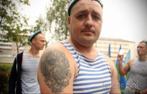 Фото тату на плече мужчины: орёл, держащий в когтях десантный шлем, с надписью, выполненное в черно-серых тонах. Детализированный рисунок татуировки с военной символикой.