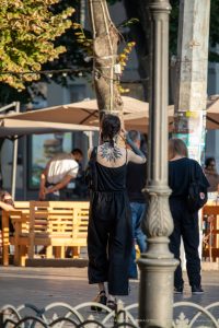 Фото тату на спине девушки в черном комбинезоне, идущей по улице. Рисунок татуировки представляет собой стилизованное изображение цветка.