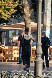 Фото тату на спине девушки в черном комбинезоне, идущей по улице. Рисунок татуировки представляет собой стилизованное изображение цветка.