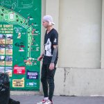 Фотография мужчины с татуировками на руках и торсе, стоящего на фоне городской стены с картой и туристической информацией.