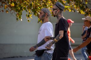 Фотография двух мужчин с обширными татуировками на шее, руках и торсе, идущих по улице в солнечный день.