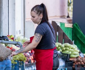 Фотография продавца на рынке с татуировкой на руке, выбирающего фрукты. Виден фрагмент черно-серого рисунка татуировки на предплечье.