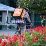 Фото тату: мужчина с татуировками на руках едет на велосипеде по улице. Видны рисунки на плече и предплечье, татуировки в стиле олдскул и традиционные.