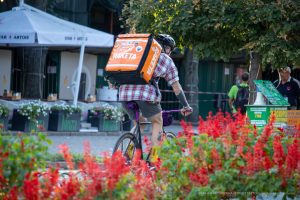 Фото тату: мужчина с татуировками на руках едет на велосипеде по улице. Видны рисунки на плече и предплечье, татуировки в стиле олдскул и традиционные.