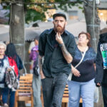 Фото тату на руке мужчины с бородой, одетого в черную рубашку и кепку, идущего по улице.