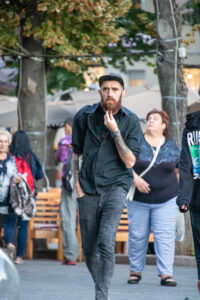 Фото тату на руке мужчины с бородой, одетого в черную рубашку и кепку, идущего по улице.