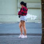 Фотография девушки, идущей по улице с видимой татуировкой на бедре. Она одета в шорты и клетчатую рубашку.