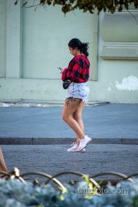 Фотография девушки, идущей по улице с видимой татуировкой на бедре. Она одета в шорты и клетчатую рубашку.