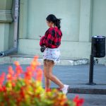 Фотография девушки, идущей по улице с видимой татуировкой на бедре. Она одета в шорты и клетчатую рубашку.