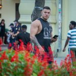 Фотография мужчины с татуировкой на руке, стоящего на улице. На заднем плане видны люди, деревья и здания.
