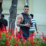 Фотография мужчины с татуировкой на руке, стоящего на улице. На заднем плане видны люди, деревья и здания.