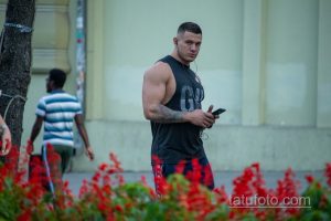 Фотография мужчины с татуировкой на руке, смотрящего в сторону. На заднем плане видны люди и цветы.