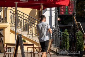 Фотография мужчины с татуировкой на руке, стоящего на улице. На заднем плане видны кафе, люди и рекламный щит с курсами валют.