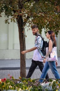 Фотография пары с татуировками на руках, гуляющей по городу под сенью деревьев. Татуировки на руках мужчины и женщины выглядят эффектно и привлекают внимание.